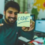 A smiling programmer pointing to a sticky note with 'code' written on it in an office setting.
