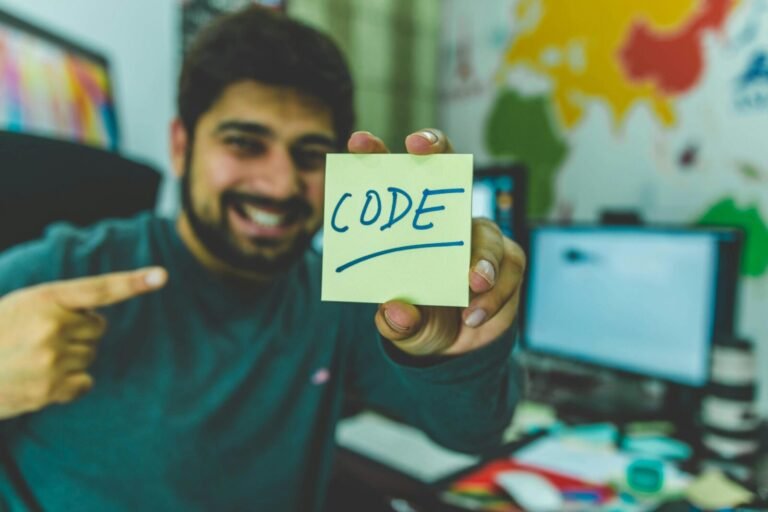 A smiling programmer pointing to a sticky note with 'code' written on it in an office setting.
